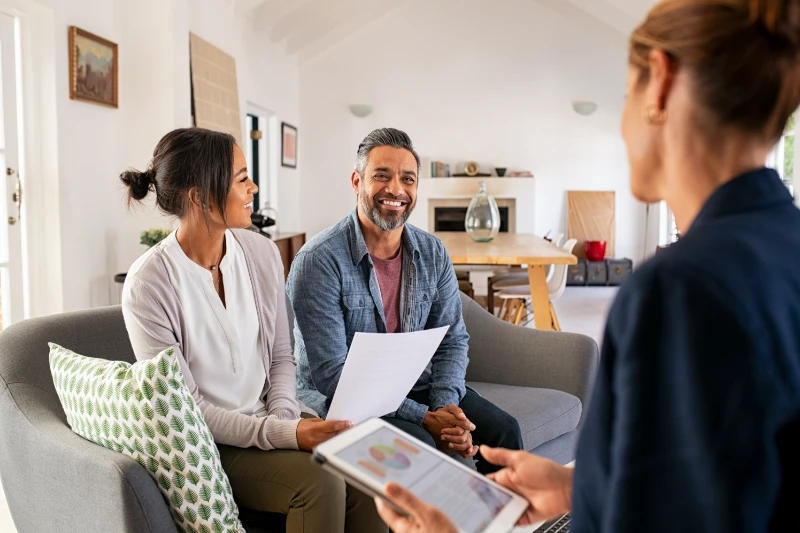 A smiling couple sits on a couch holding documents while talking to a professional woman with a digital tablet in a bright, modern living room.